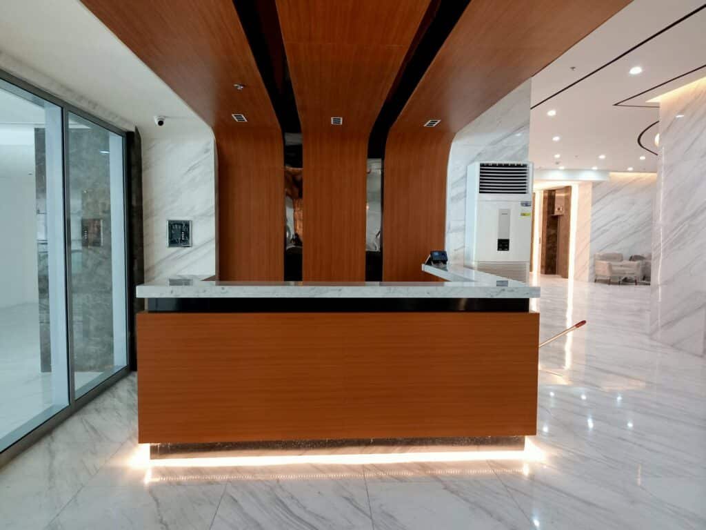 Modern reception desk with wood paneling and marble countertop in a brightly lit office lobby with white marble floors and walls.