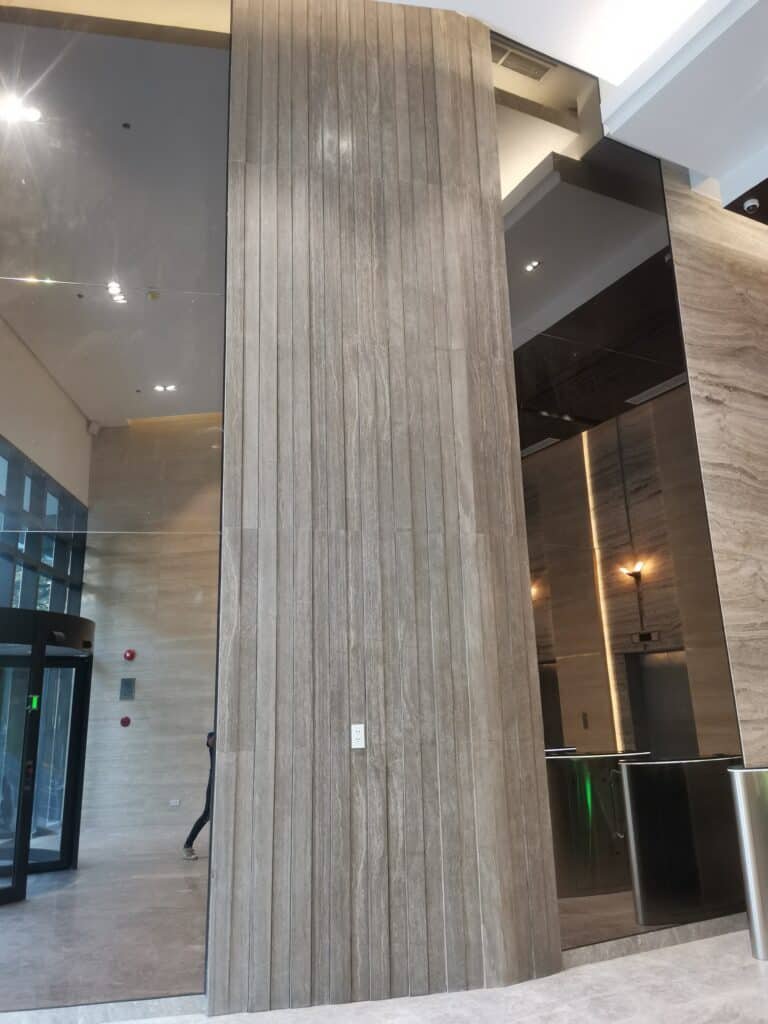 Tall, modern lobby wall with vertical wooden panels and a single white switch; glass doors and reflective surfaces visible on both sides.