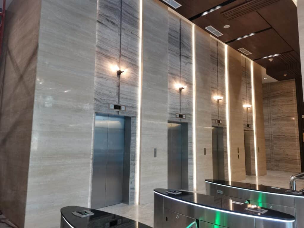Four closed elevator doors in a modern building lobby with marble walls, vertical lights, and electronic access gates in the foreground.