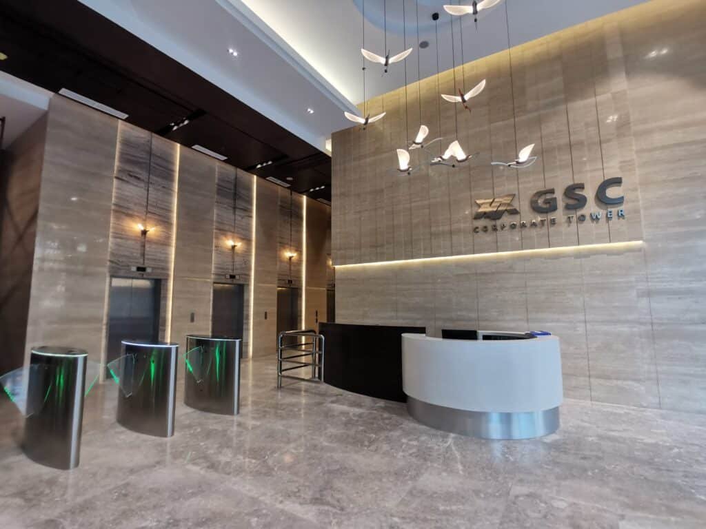 Modern office building lobby with turnstiles, a curved reception desk, elevators, and "GSC Corporate Tower" signage on a marble wall. Contemporary lighting fixtures hang from the ceiling.