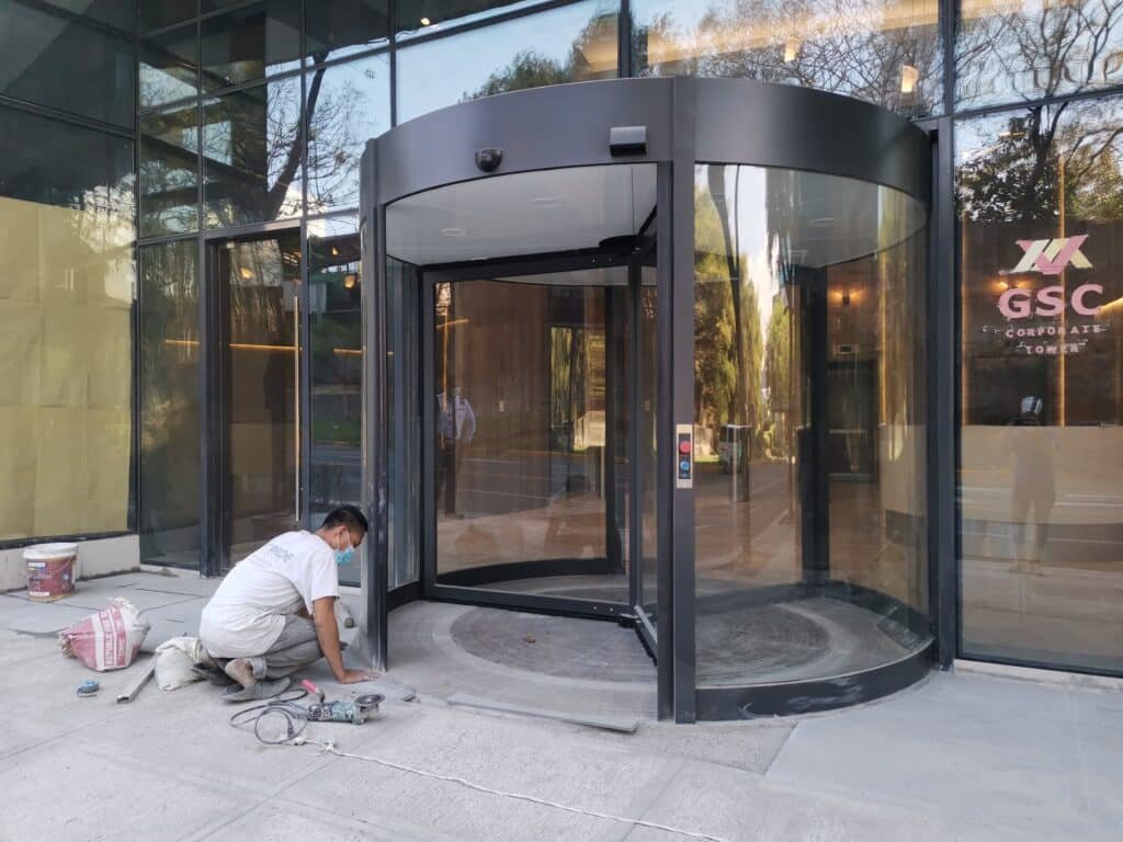 A worker kneels on the ground installing or repairing equipment near a large glass revolving door at the entrance of a modern building.