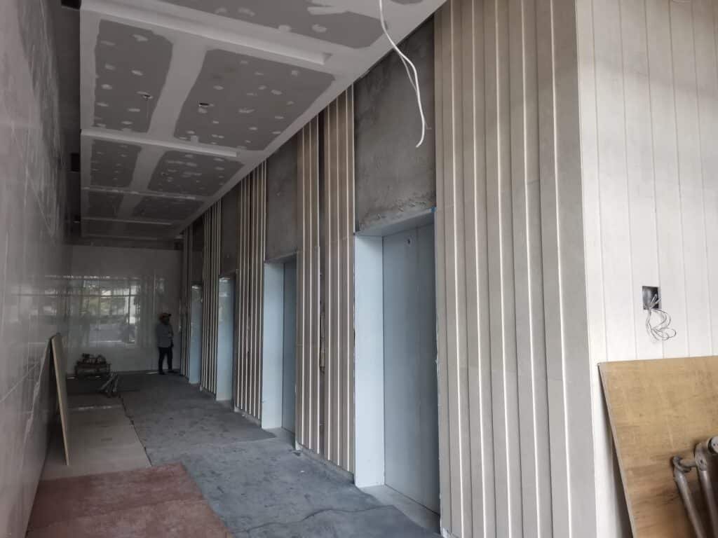 An unfinished interior corridor with exposed elevator doors, unpainted drywall ceiling, construction materials, and a person standing in the background.