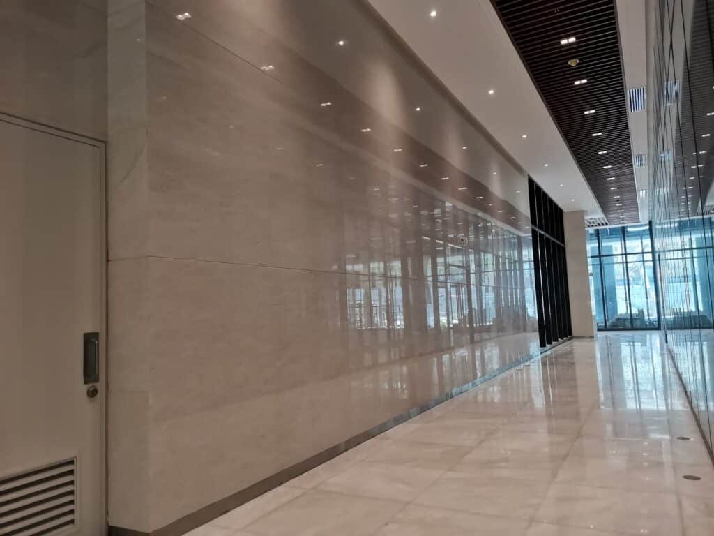 Wide, empty hallway with polished marble floors, beige walls, overhead lights, and large windows letting in natural light at the far end.