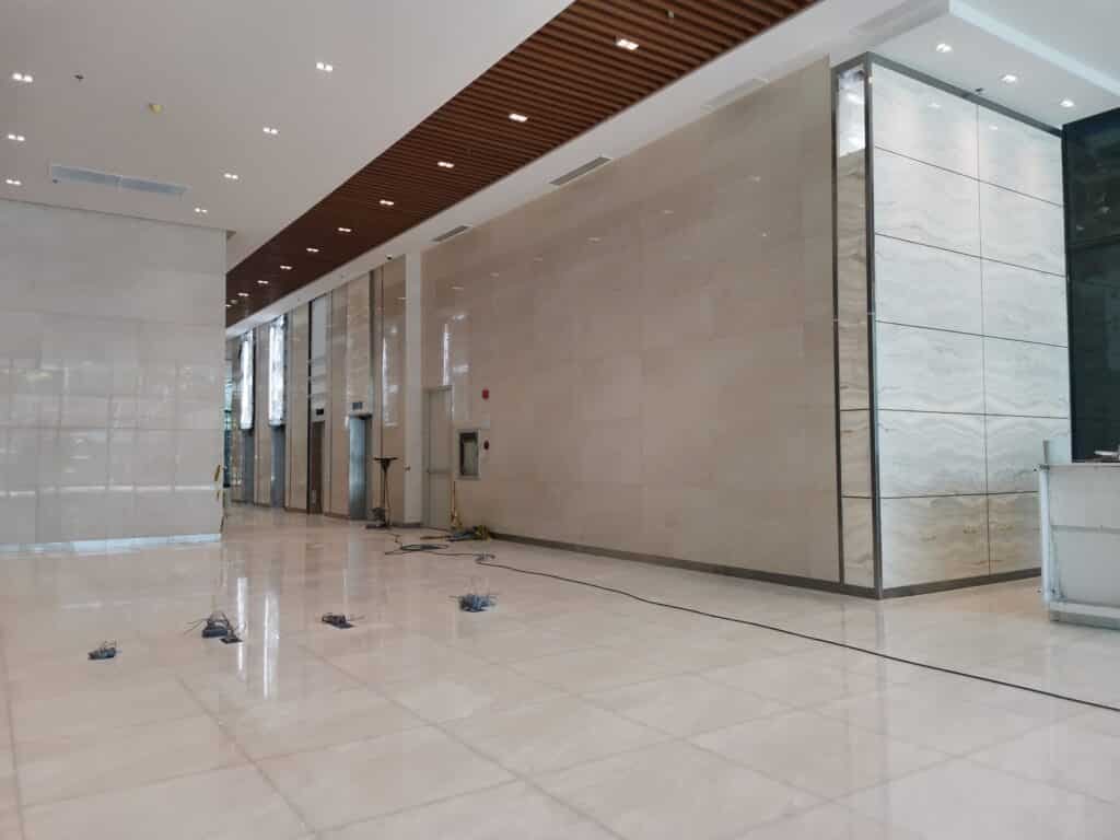 A spacious, empty lobby with shiny tiled floors, marble walls, a wooden slat ceiling, visible wiring, and some equipment on the floor.