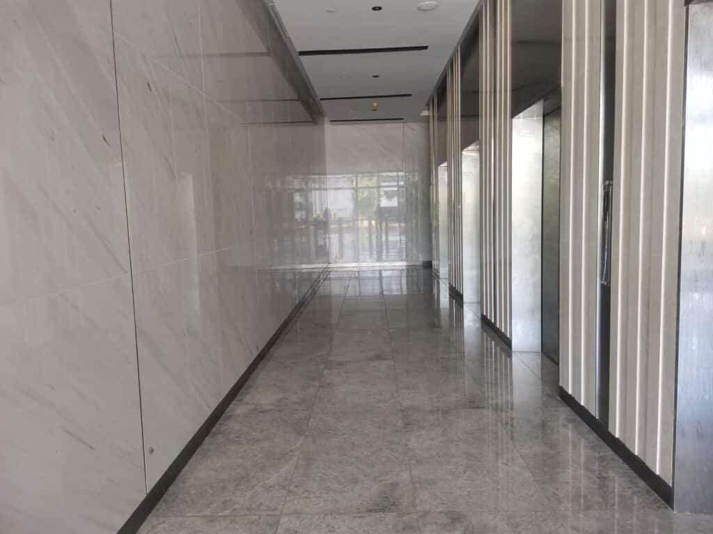 A modern hallway with marble walls and floors, reflecting light from glass doors at the end, and elevator doors on the right side.