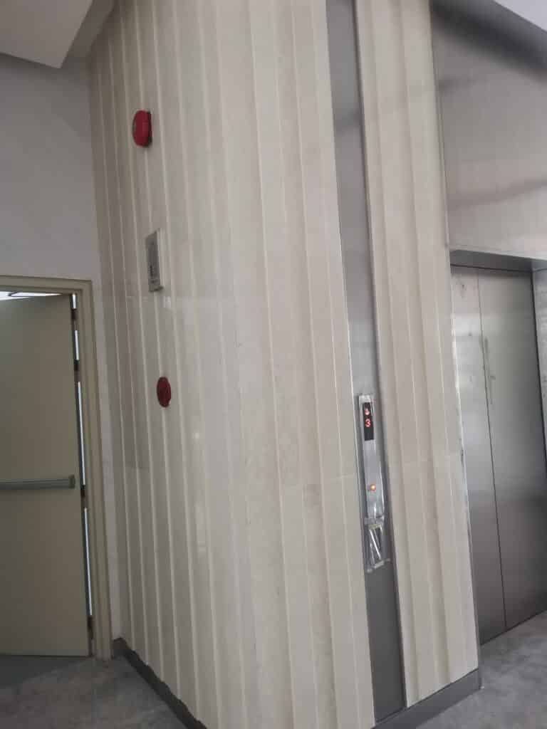 A closed elevator with a control panel showing the number 3, located next to a partially open door and two red emergency alarms on a marble wall.