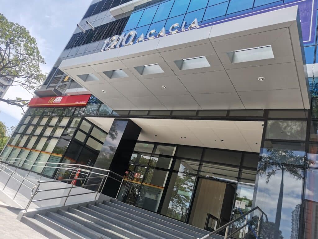 Modern building entrance with a ramp and stairs, glass facade, and signage reading "1210 ACACIA;" bank branch sign visible on the left.