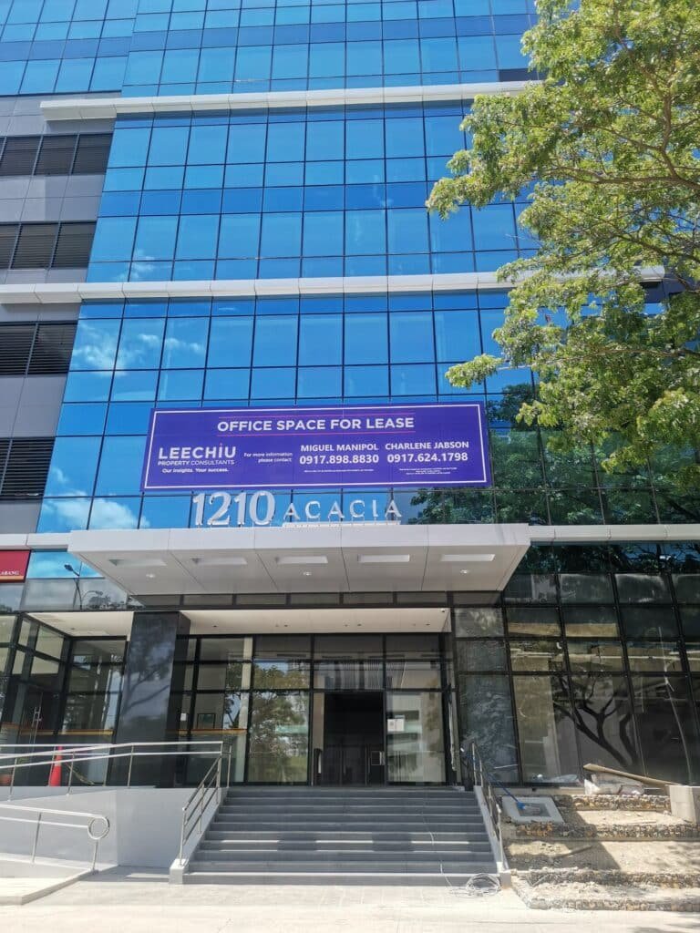 A modern glass office building with a sign above the entrance that reads "Office Space For Lease" along with contact information. The address "1210 Acacia" is visible.