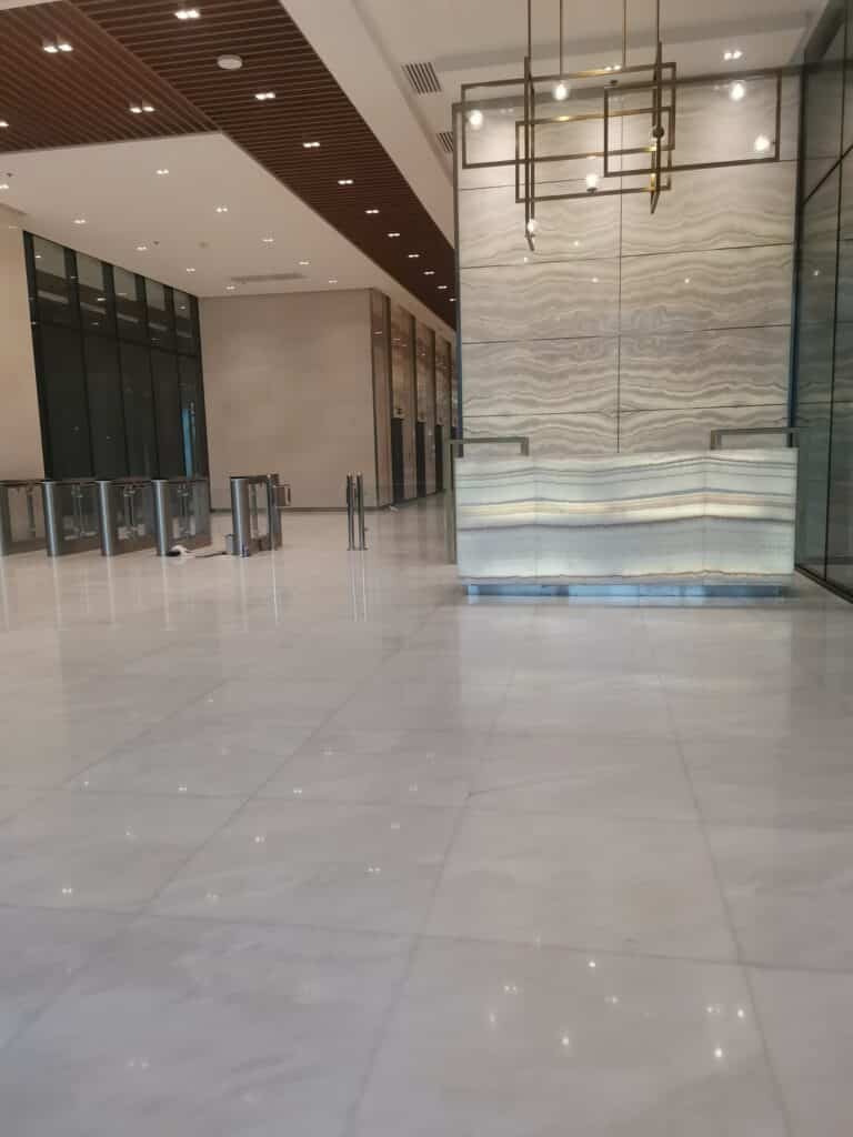 Spacious modern lobby with a marble floor, glass entrance barriers, and a reception desk with stone paneling under bright ceiling lights.