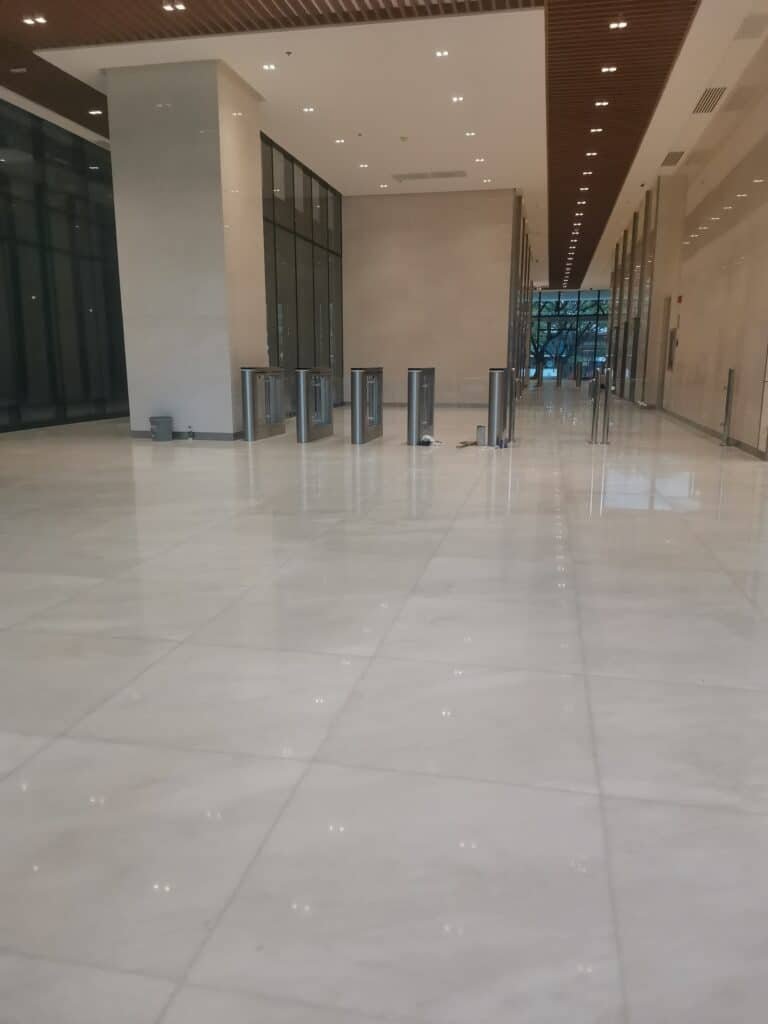 A spacious, empty building lobby with shiny tiled floors, glass walls, and security turnstiles near the entrance.