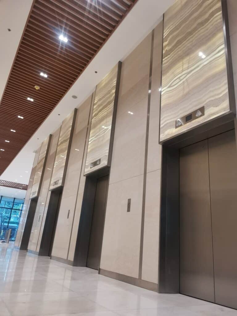 A modern building interior with four closed stainless steel elevator doors set in marble walls, under a high ceiling with wooden slats.