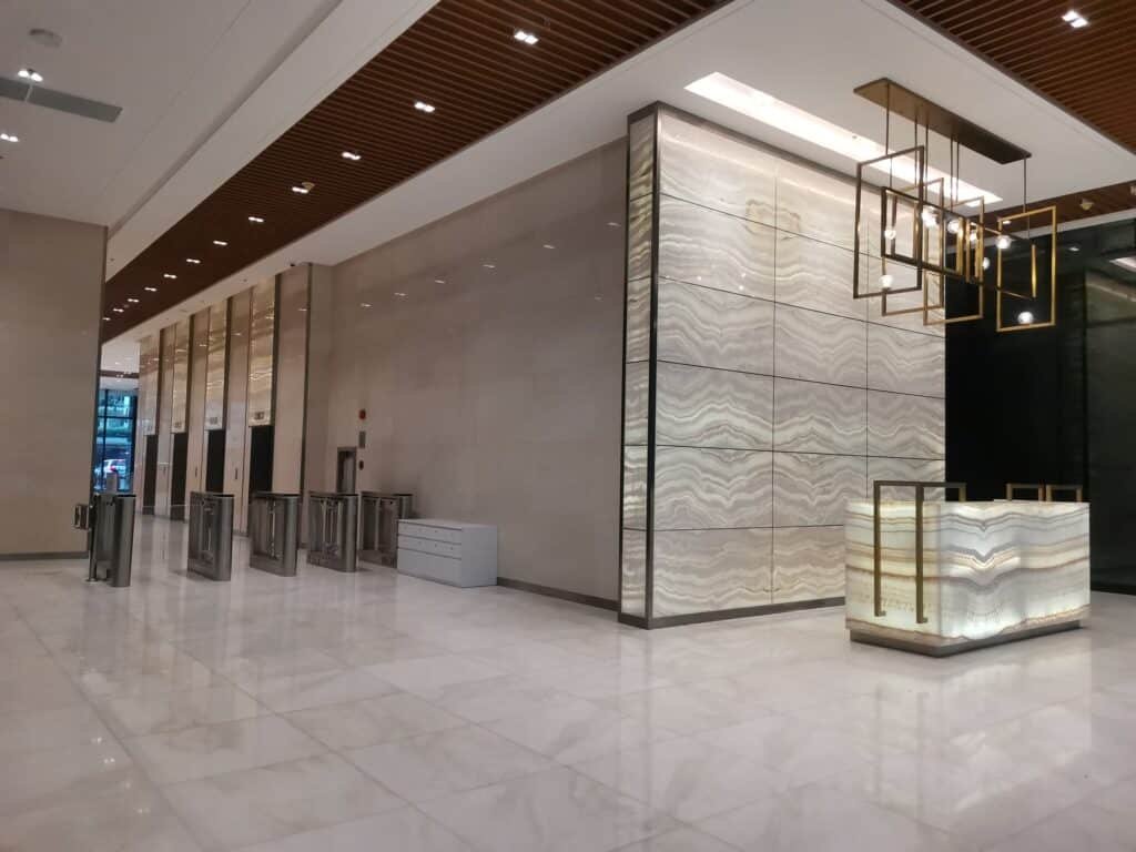 Spacious modern lobby with polished marble floors, geometric light fixtures, a reception desk, and security turnstiles along the left wall.