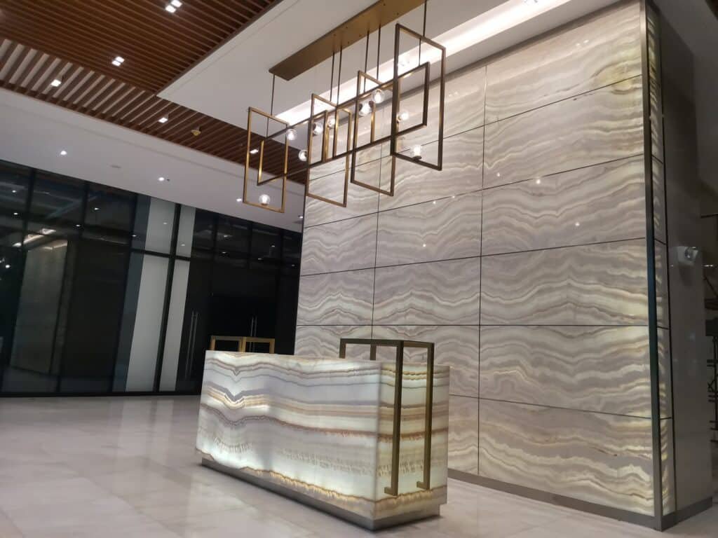 Modern hotel lobby with a marble reception desk, illuminated ceiling lights, and a large marble feature wall. Floor and walls are light-colored with a geometric ceiling design.