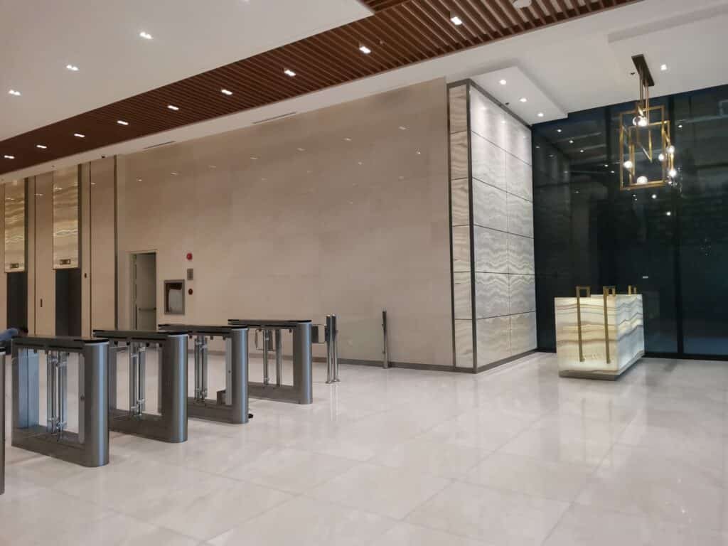 Modern office lobby with security turnstiles, marble floors, beige walls, a reception desk, and contemporary lighting fixtures.