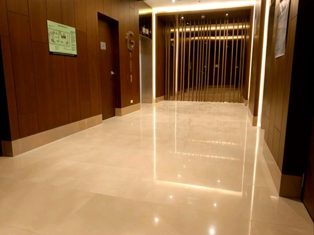 A well-lit hallway with polished beige floors, brown wood-paneled walls, and doors on the left side; vertical slats are visible at the end.