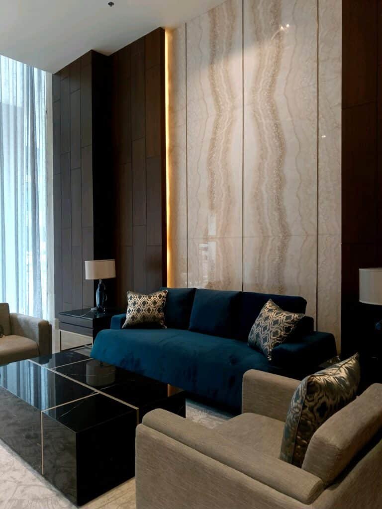 Modern living room with a blue sofa, two beige armchairs, patterned cushions, marble accent wall, black coffee tables, and two lamps on dark side tables.