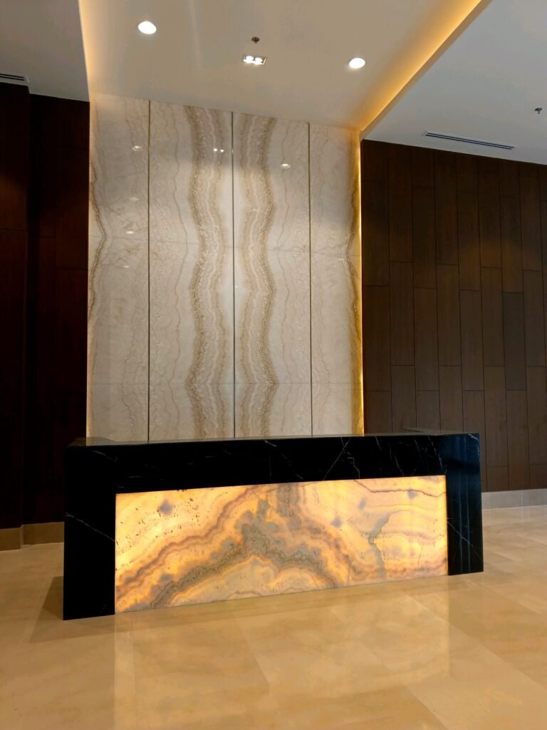 Modern reception desk with a backlit marble front panel and marble wall backdrop, in a minimalist lobby with beige flooring and wood-paneled walls.