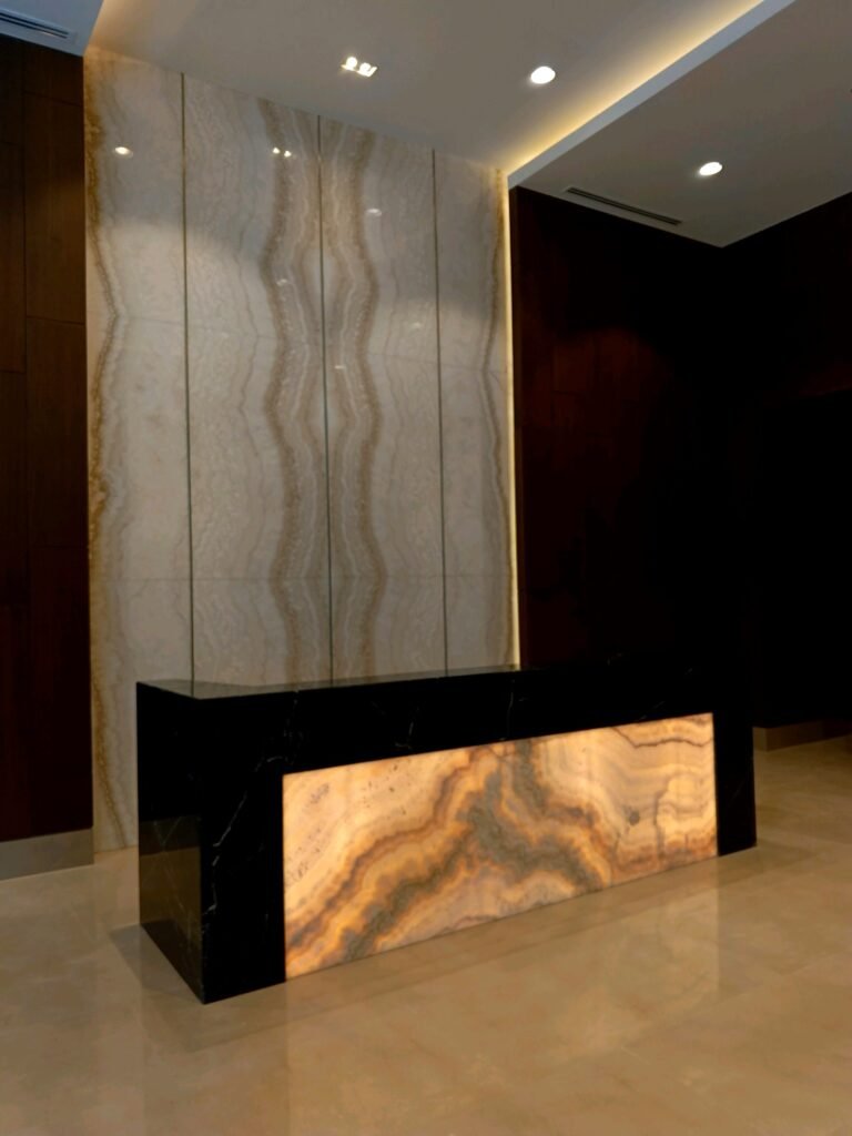 A modern reception desk with a backlit onyx front panel stands before a large polished stone wall in a dimly lit lobby.