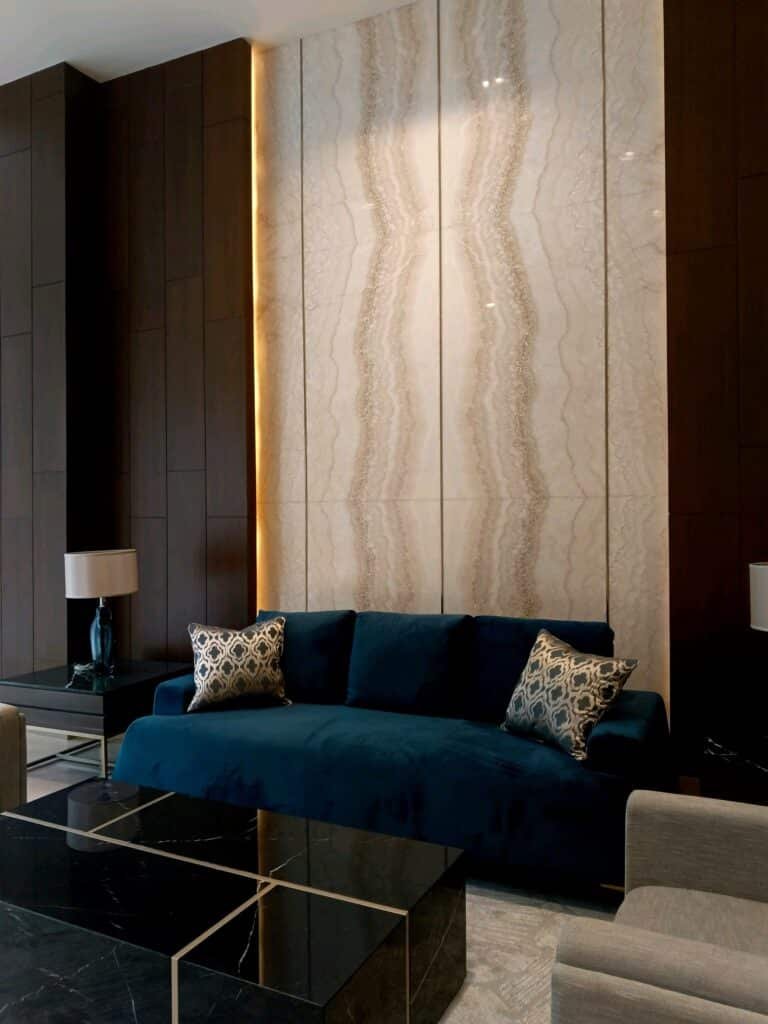 A modern living room with a dark blue sofa, patterned pillows, marble wall panels, side tables with lamps, and a black marble coffee table.