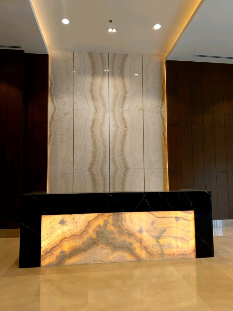 A modern reception desk with a backlit onyx front panel and a marble wall backdrop in a lobby with neutral tones.