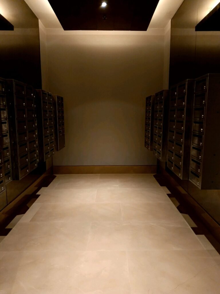 A dimly lit room with rows of metal mailboxes mounted on both side walls, and a tiled floor leading to a blank back wall.