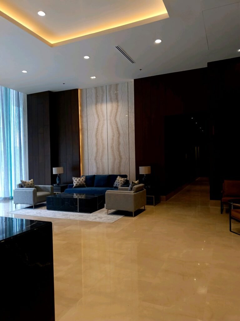 Modern hotel lobby with polished marble floors, blue and gray sofas, a marble accent wall, and ambient ceiling lighting.