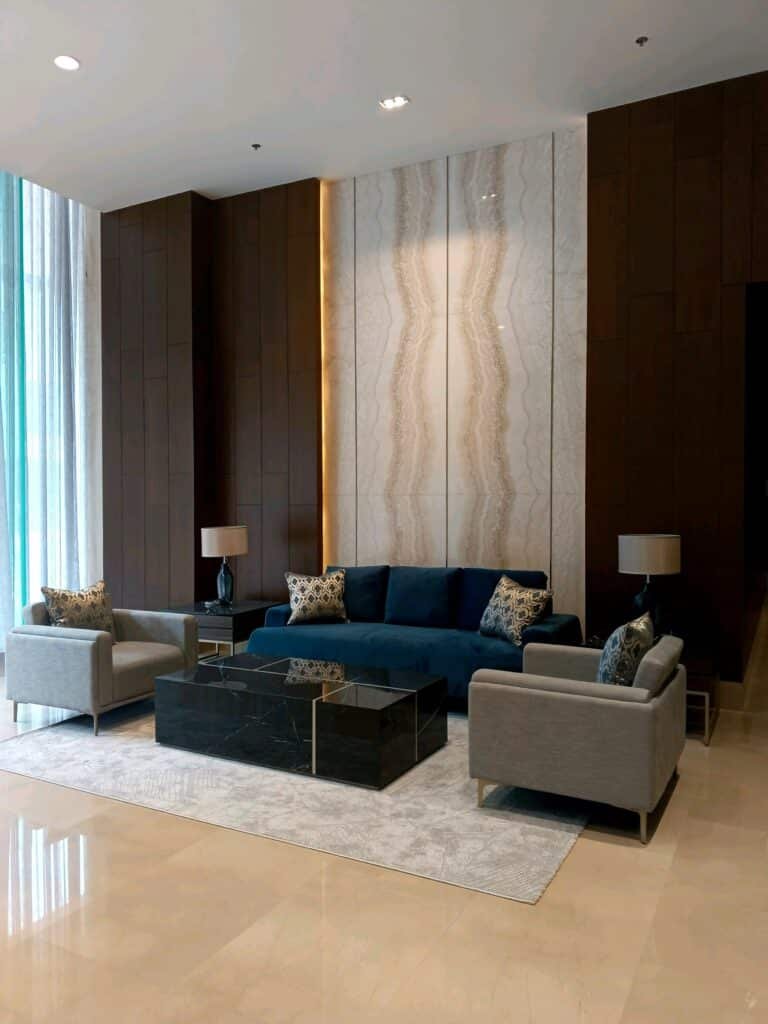 Modern lobby seating area with a blue sofa, two grey armchairs, a glass coffee table, two table lamps, and decorative pillows on a light rug against a marble accent wall.