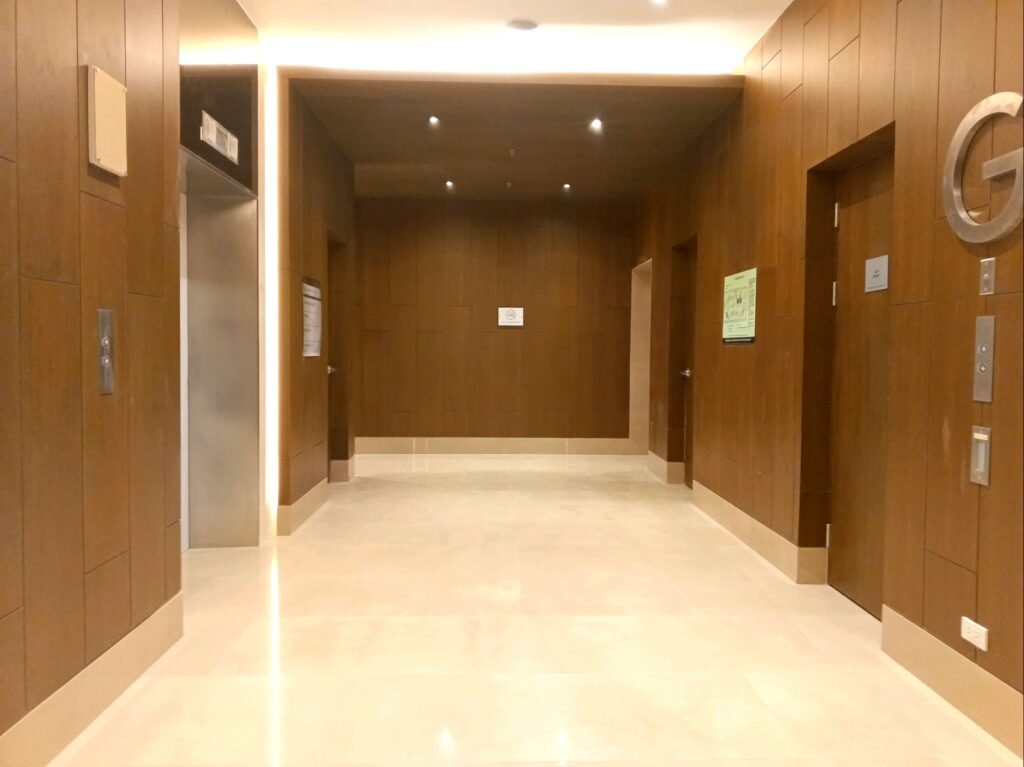 A well-lit hallway with polished floors and four closed elevator doors set in wood-paneled walls, with signs and buttons next to each elevator.