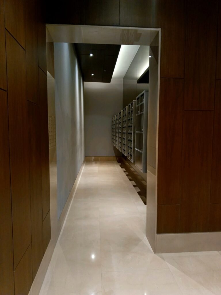 A hallway with beige tile floors and wood-paneled walls leads to a room containing multiple rows of silver mailboxes on the right side.