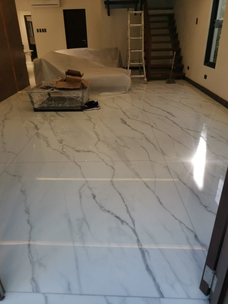 A room with white marble flooring, covered furniture, a step ladder by wooden stairs, and some boxes and shoes on the floor.