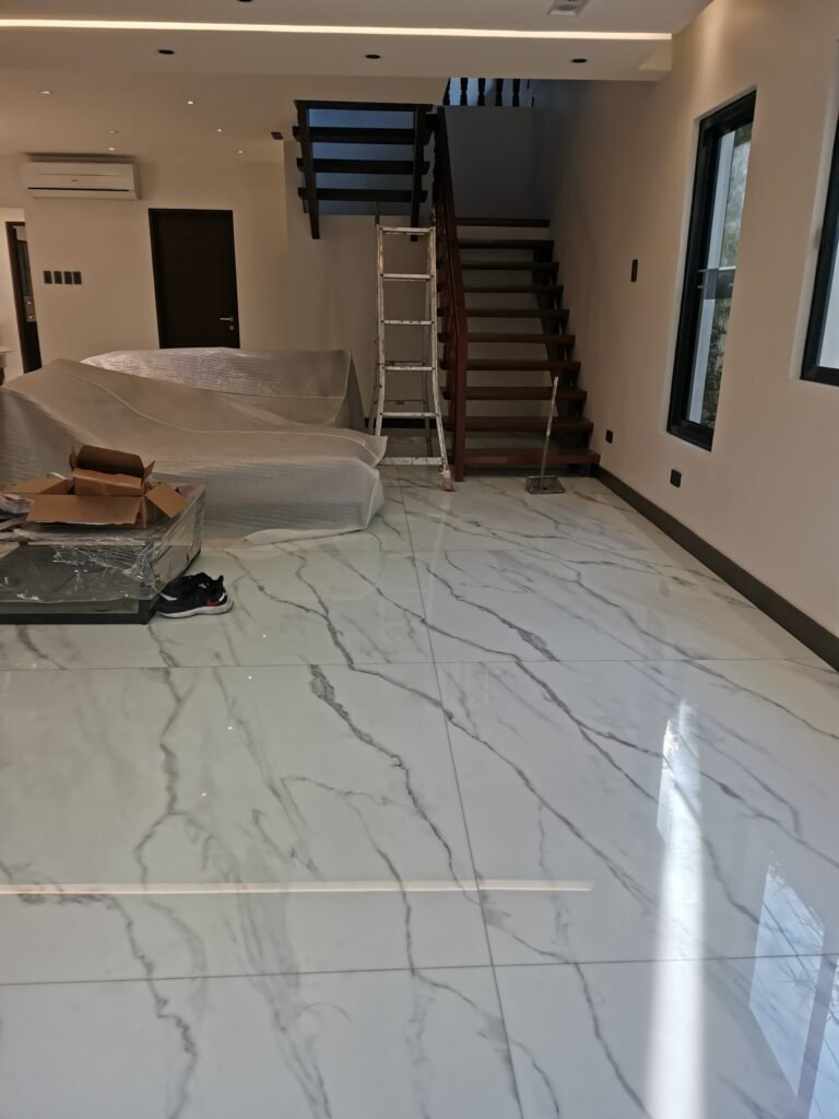 Modern interior with glossy marble floor, covered furniture, boxes, and a ladder by a wooden staircase, suggesting ongoing work or recent move-in.