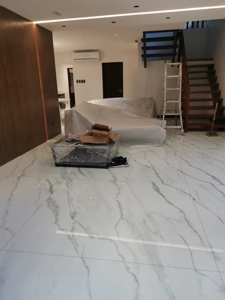 A modern living room under renovation with covered furniture, a ladder by the staircase, and boxes on a marble floor.