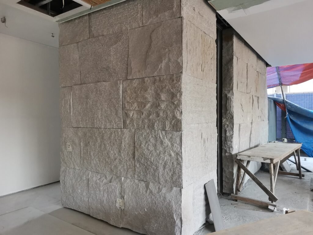 A stone-clad rectangular structure under construction indoors, with unfinished walls, exposed ceiling, and construction materials nearby.