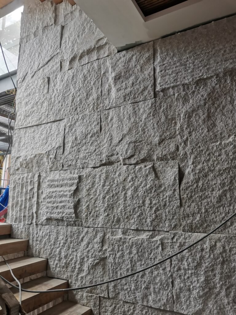 A stone wall with large, rough rectangular blocks sits beside a wooden staircase in an unfinished indoor construction area.