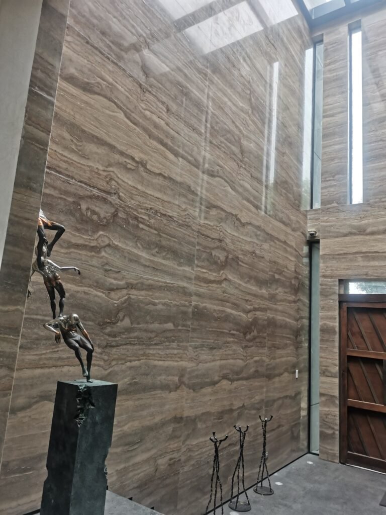 Modern foyer with tall, brown marble walls, a skylight, a wooden door, a sculpture of figures stacked on a pedestal, and several small metal figurines on the floor.