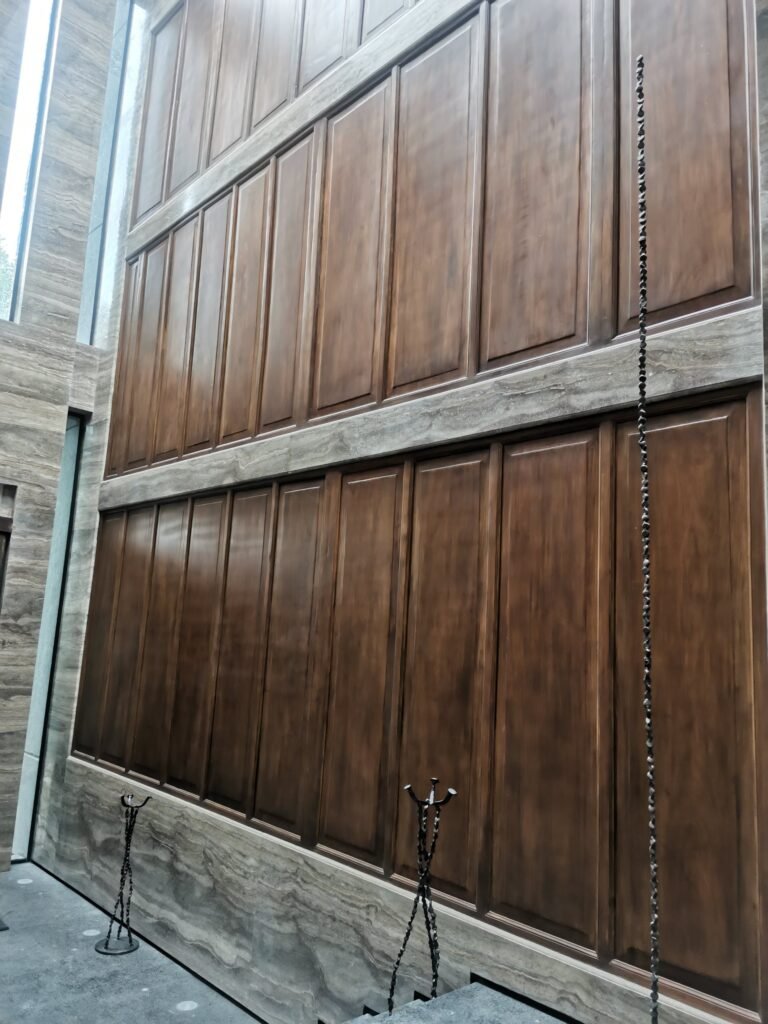 Tall wooden paneled wall with vertical windows on the left and metal candle holders in front, set atop stone flooring and walls.