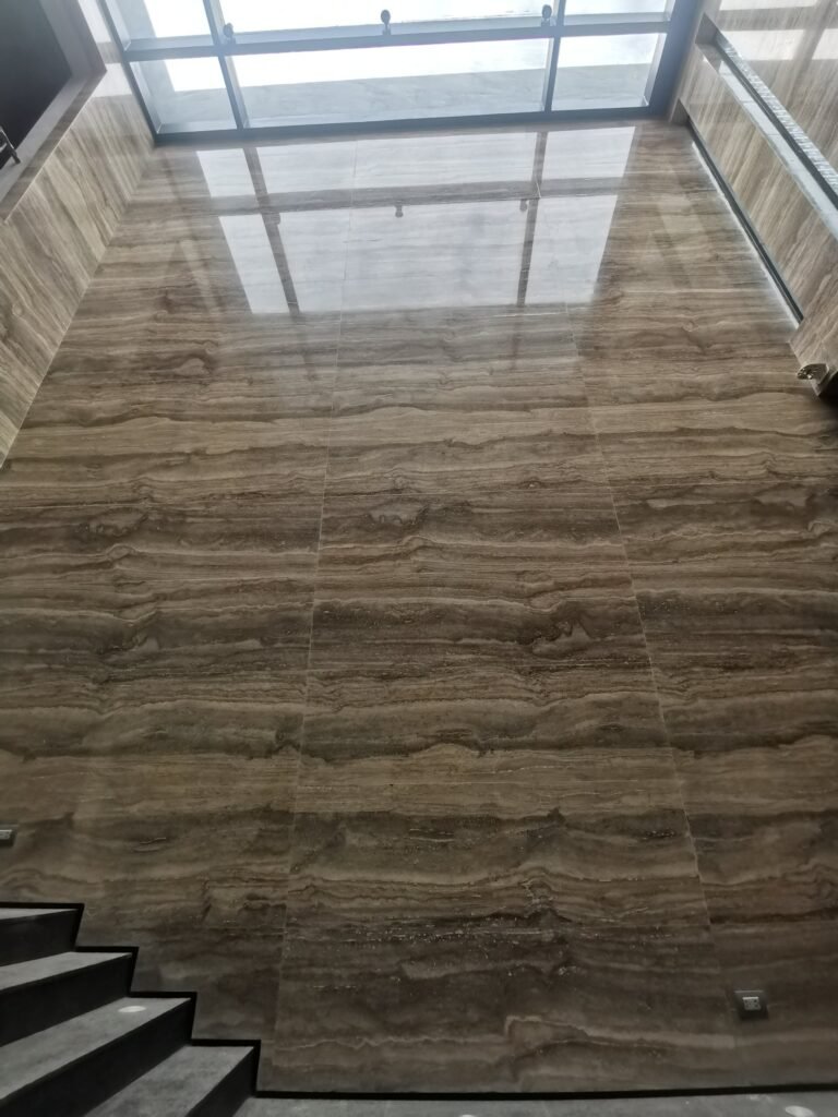 Large marble wall with horizontal brown and beige veins, viewed from a staircase below, with a glass ceiling or skylight at the top letting in natural light.