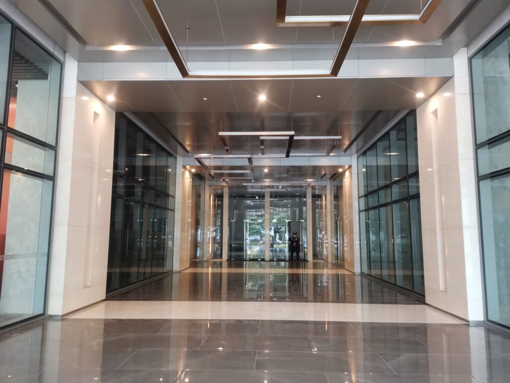 A modern building corridor with glass walls, polished floors, and ceiling lights, leading to an entrance with trees visible outside.