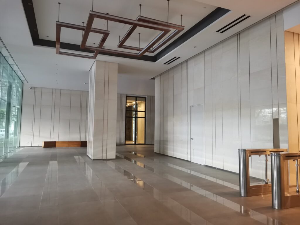 Spacious modern building lobby with large tiled floor, tall columns, glass wall on the left, wooden bench at the back, and metal turnstiles in the foreground.