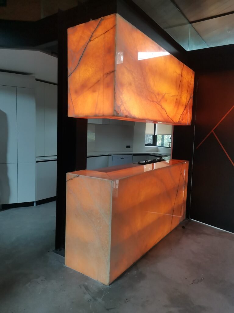 A modern kitchen features a rectangular bar counter and matching overhead unit made of orange-hued, backlit translucent stone with visible veining.