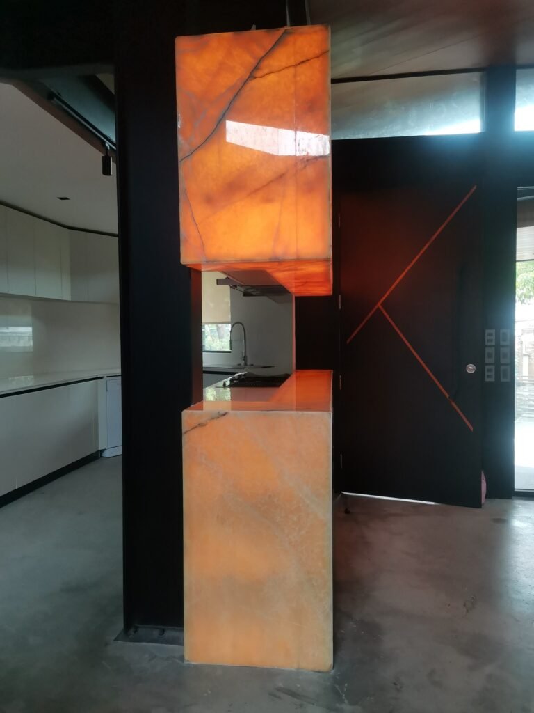Two rectangular, illuminated orange onyx panels are mounted vertically in a modern kitchen with concrete floors and black walls.