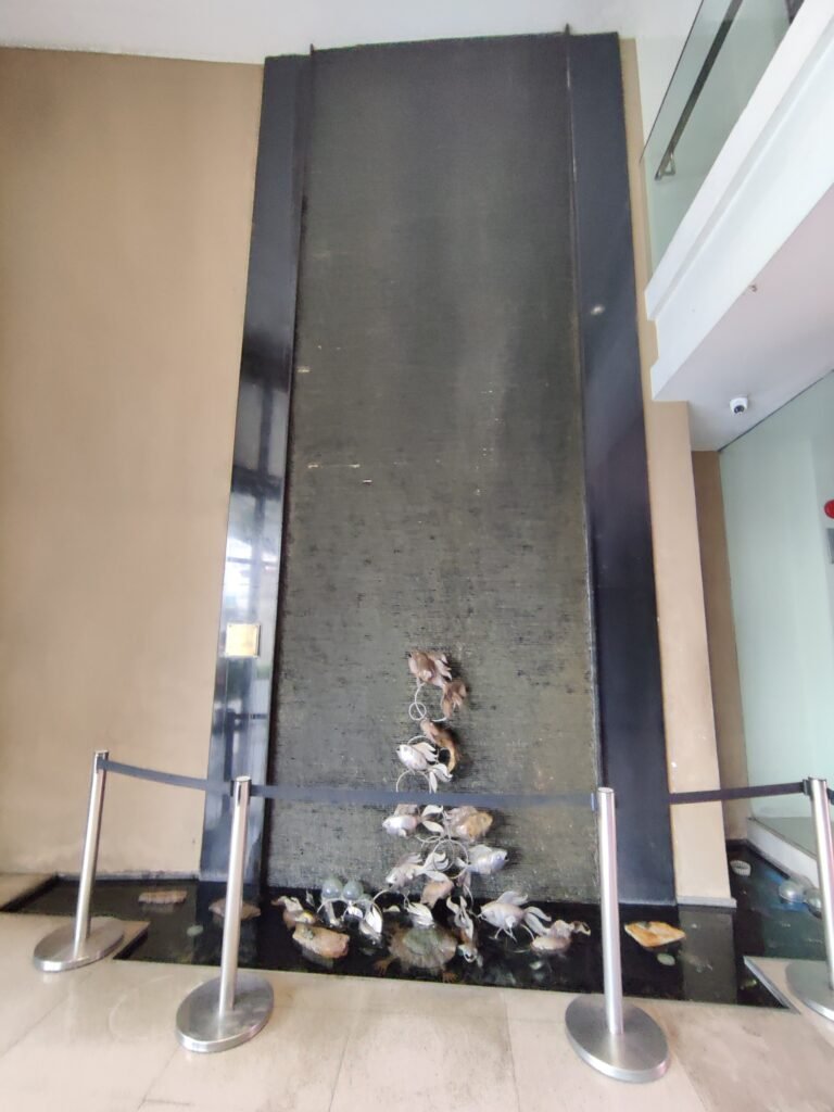 Indoor water feature with a vertical black stone wall, cascading water, and decorative rocks at the base, partially surrounded by a black rope barrier.