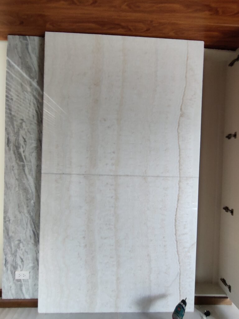 Two large stone slabs, one white with subtle beige veining and one gray with darker patterns, are displayed vertically indoors against a white wall and wood floor.