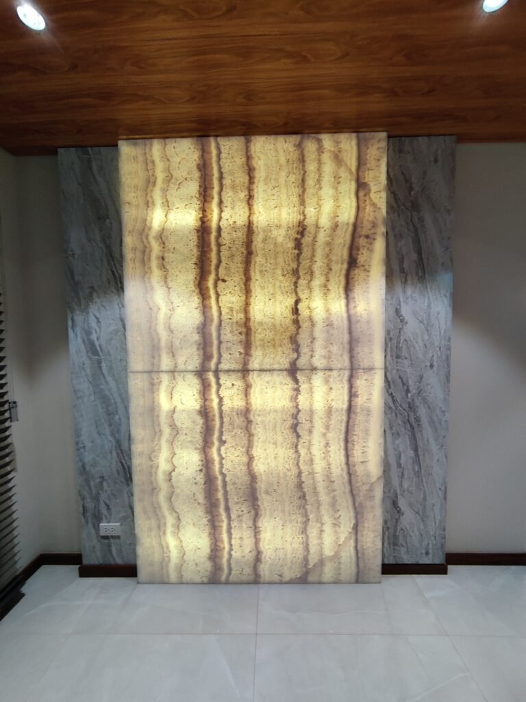 A backlit onyx stone wall panel with vertical brown stripes is mounted against a grey marble accent wall, under a wooden ceiling with recessed lights.