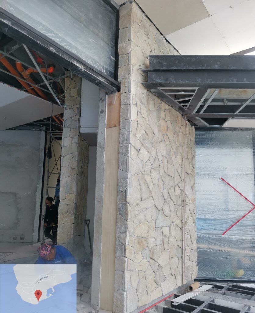 Two workers are inside a building under construction with stone veneer walls, exposed ceiling framework, and partially installed glass panels. A small map is visible in the bottom left corner.