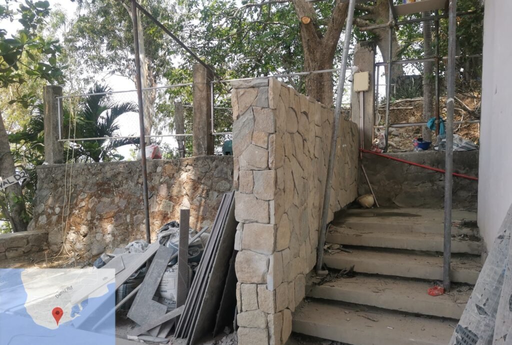 Outdoor stone staircase under construction with building materials and debris nearby; partial stone wall and trees in the background.