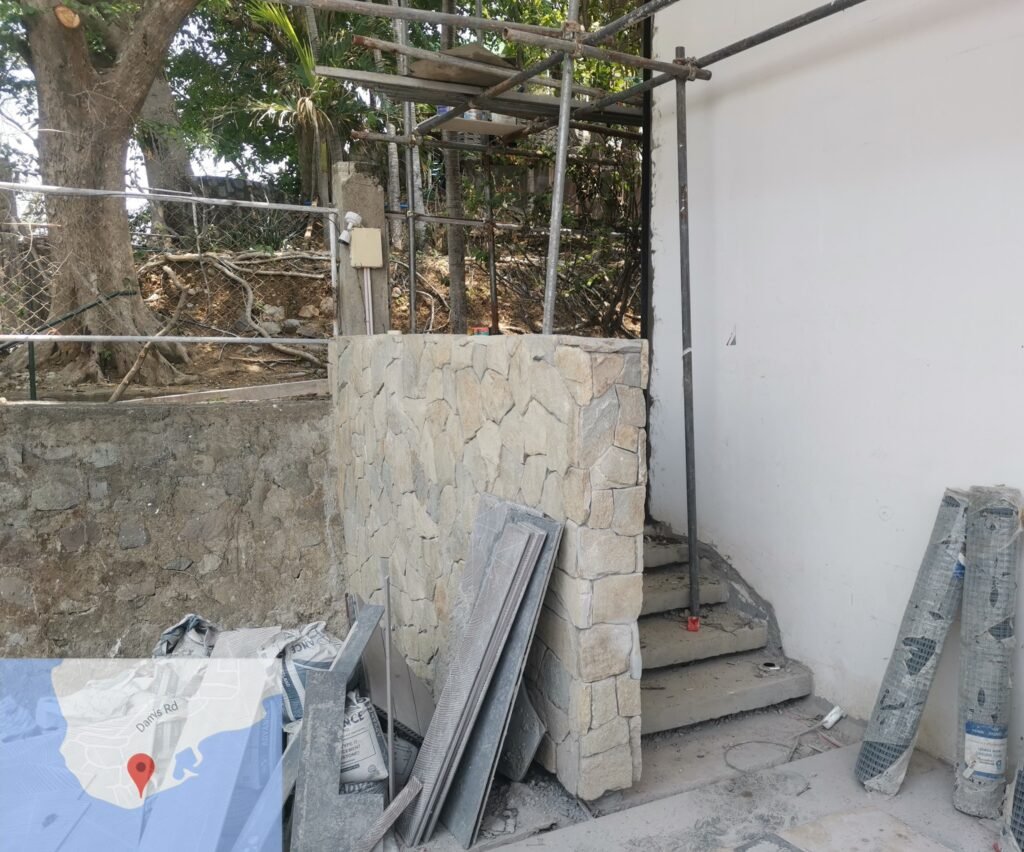 A stone wall and partially constructed staircase with scaffolding and building materials placed nearby; a tree and fence are in the background.