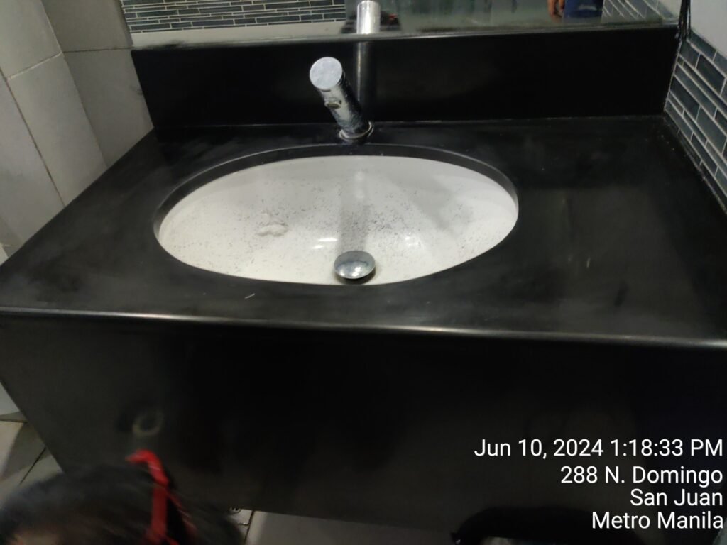 A black sink with water stains and a single faucet, set against a tiled wall and mirror. Date, time, and location text is overlaid in the bottom right corner.