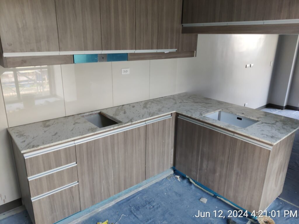 Unfinished kitchen with light wood cabinets, marble-patterned countertops, and empty spaces for appliances. Construction materials and debris are visible on the floor.