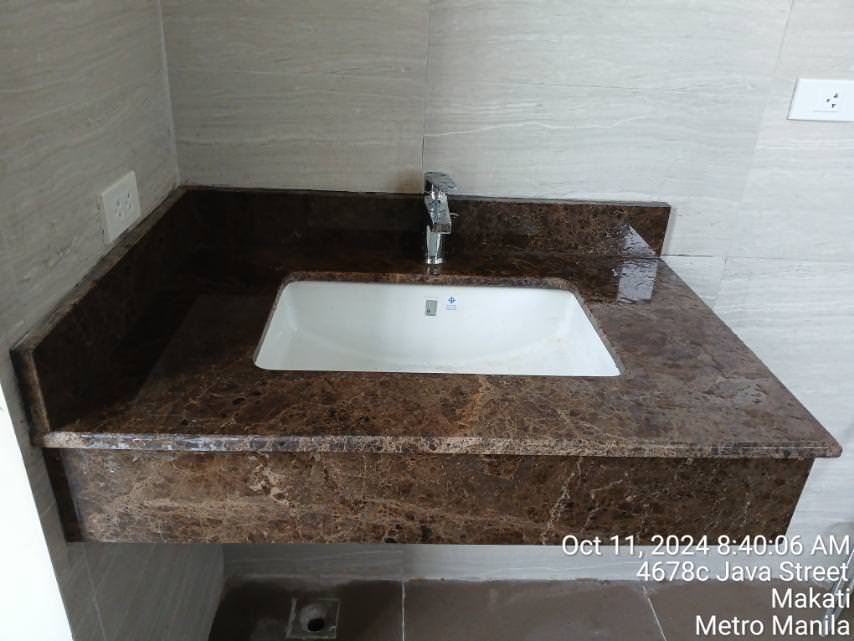 A rectangular sink with a brown marble countertop and chrome faucet, set against light-colored tiled walls. An electrical outlet is on the left wall.